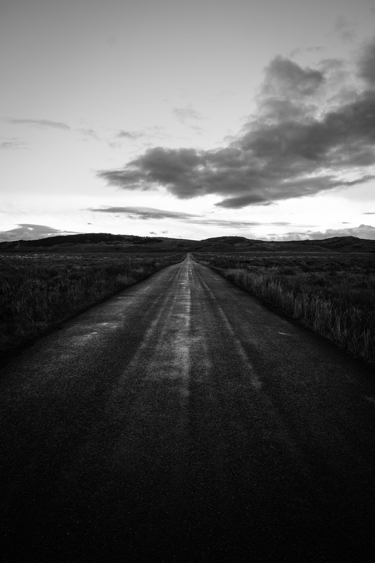 A long, straight road extending into the distance with grassy fields on either side. The horizon shows low hills and a sky filled with scattered clouds.