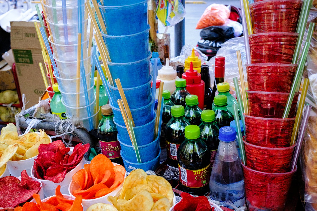 A street food stall with plastic cups, bottled drinks, various sauces, and colorful snack chips displayed.
