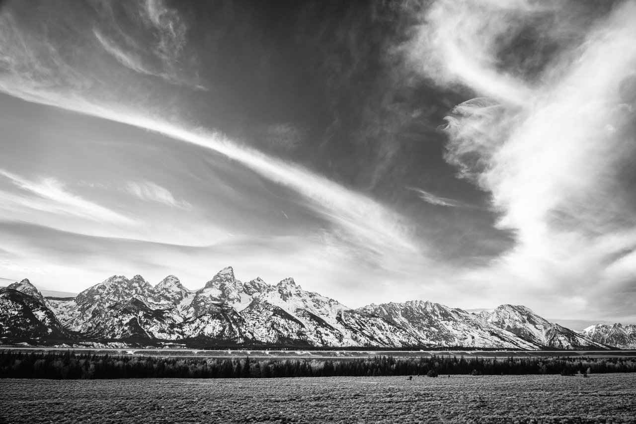 The Teton Range, with jagged, snow-covered peaks, rising above a flat valley floor with scattered vegetation and trees, beneath a sky filled with sweeping cirrus clouds forming dramatic streaked patterns.